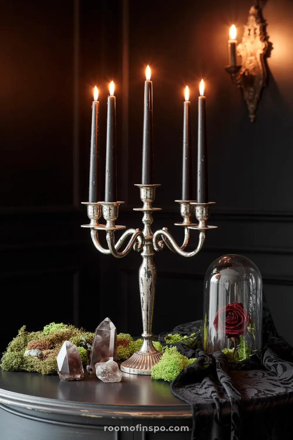 A mystical Halloween centerpiece features a silver candelabra with black candles, moss, crystals, and a single red rose under a cloche.