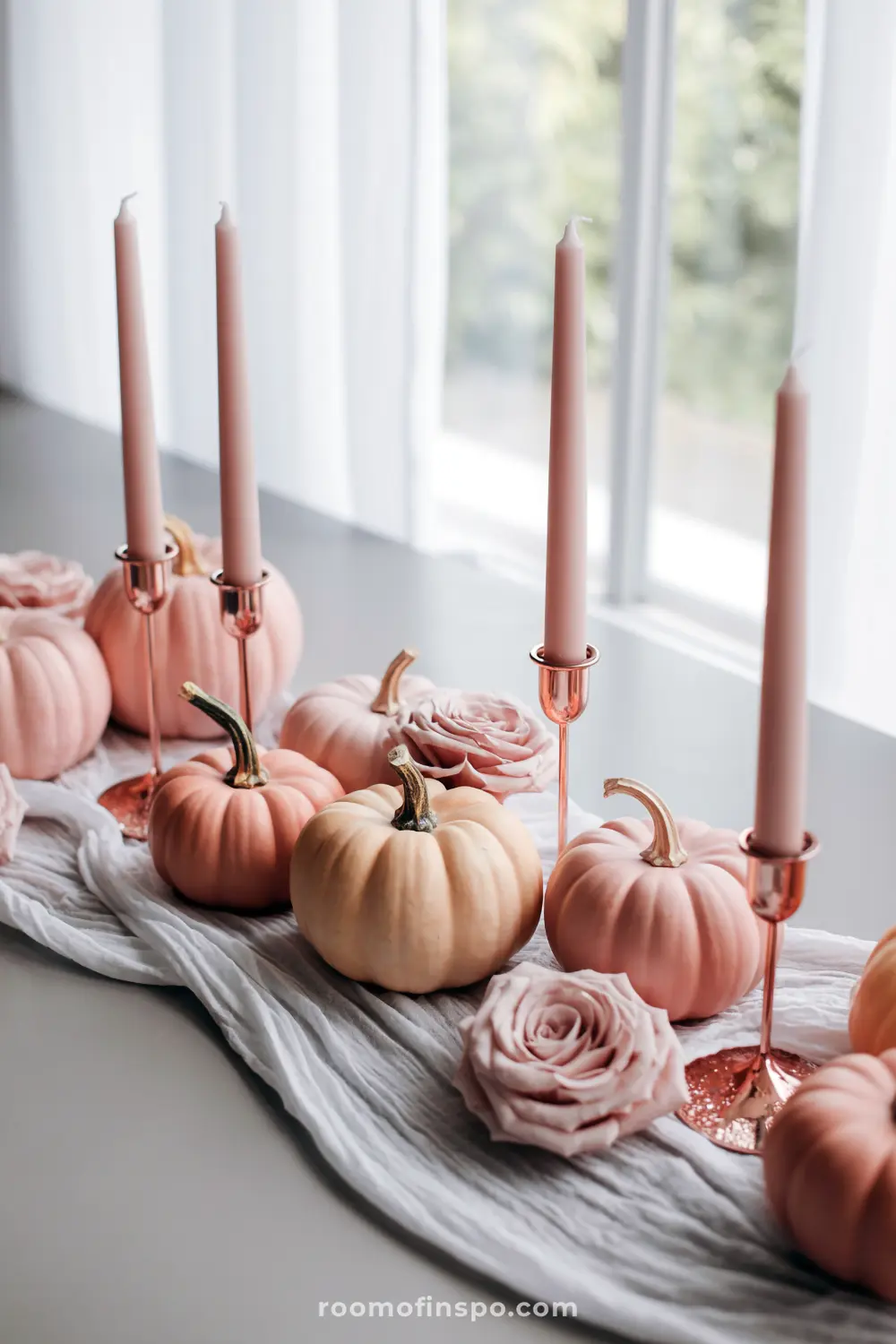 A serene Halloween centerpiece featuring a mix of pink and peach pumpkins, pink roses, and tall pink taper candles on a light runner.