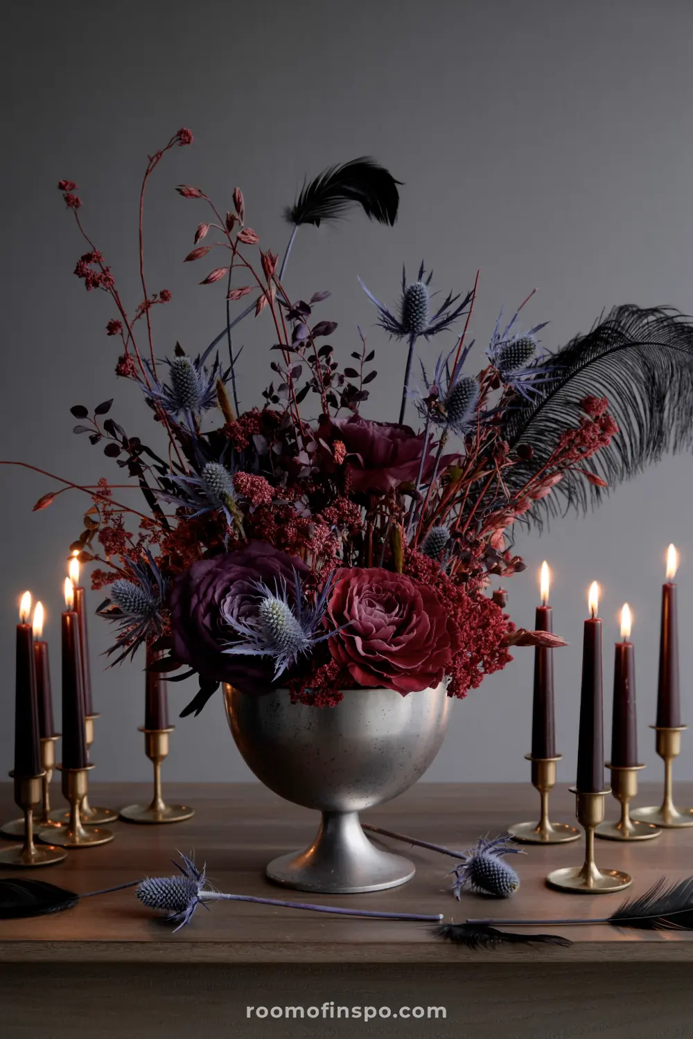 A dramatic Halloween centerpiece in a silver bowl features dark red florals, thistles, and black feathers, surrounded by burning candles.