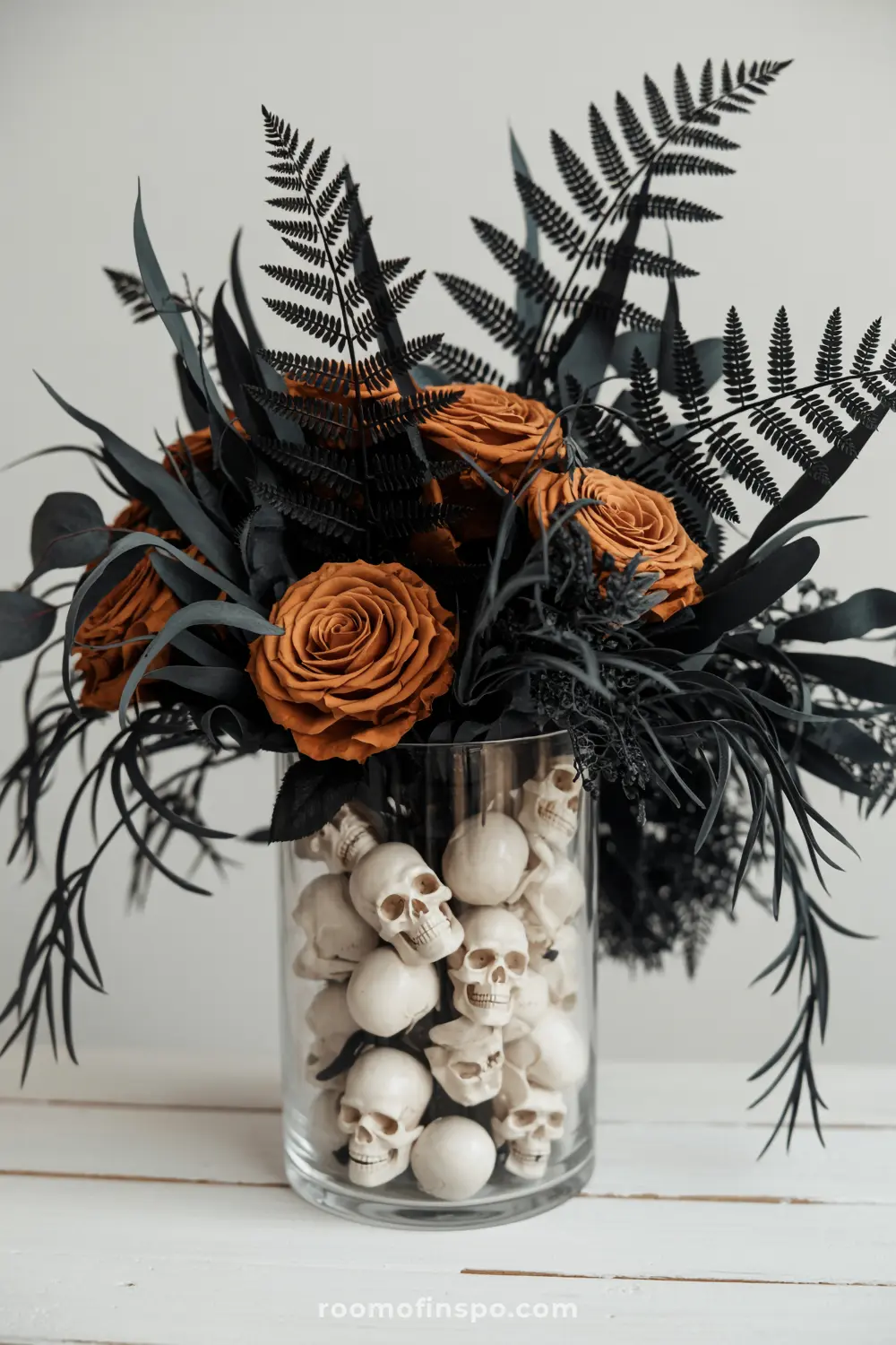 A unique Halloween centerpiece with orange roses and dark foliage emerging from a glass vase filled with miniature skulls.