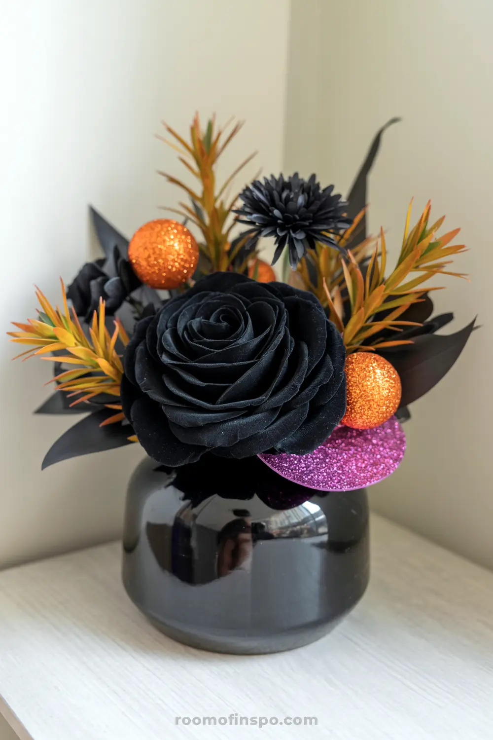 A striking Halloween centerpiece in a black vase featuring black roses, vibrant orange foliage, glittery spheres, and a purple disk.