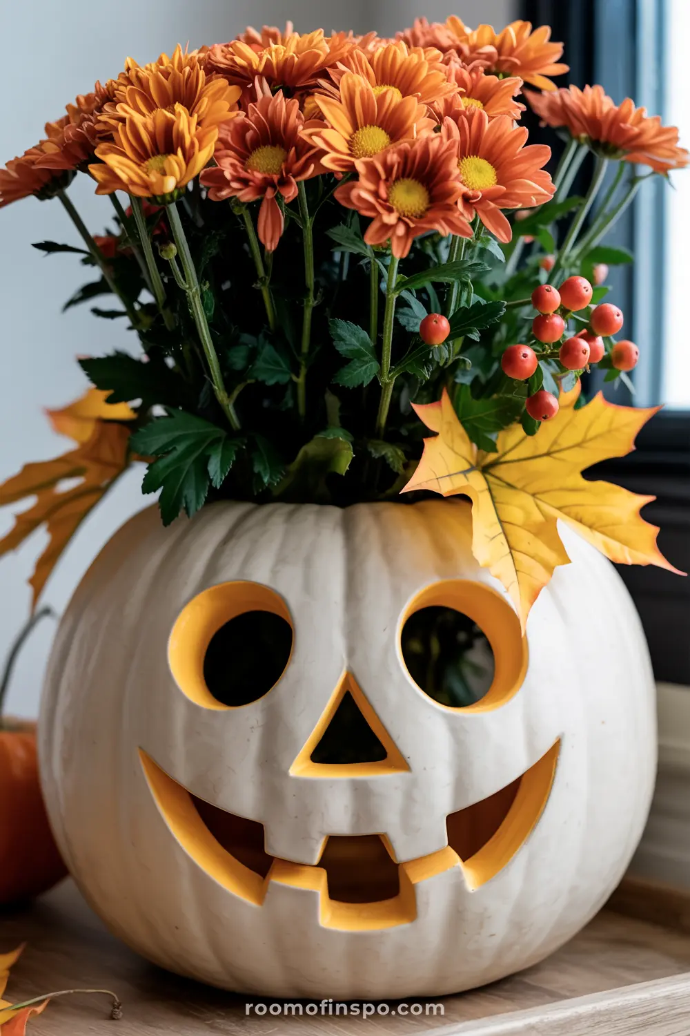 A cheerful Halloween centerpiece features a white carved pumpkin with a friendly face, filled with bright orange chrysanthemums and red berries.