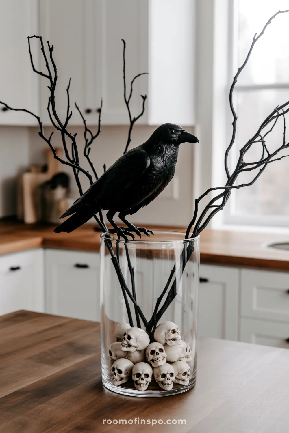A stark Halloween centerpiece features a black crow perched on bare branches emerging from a clear vase filled with small skulls.