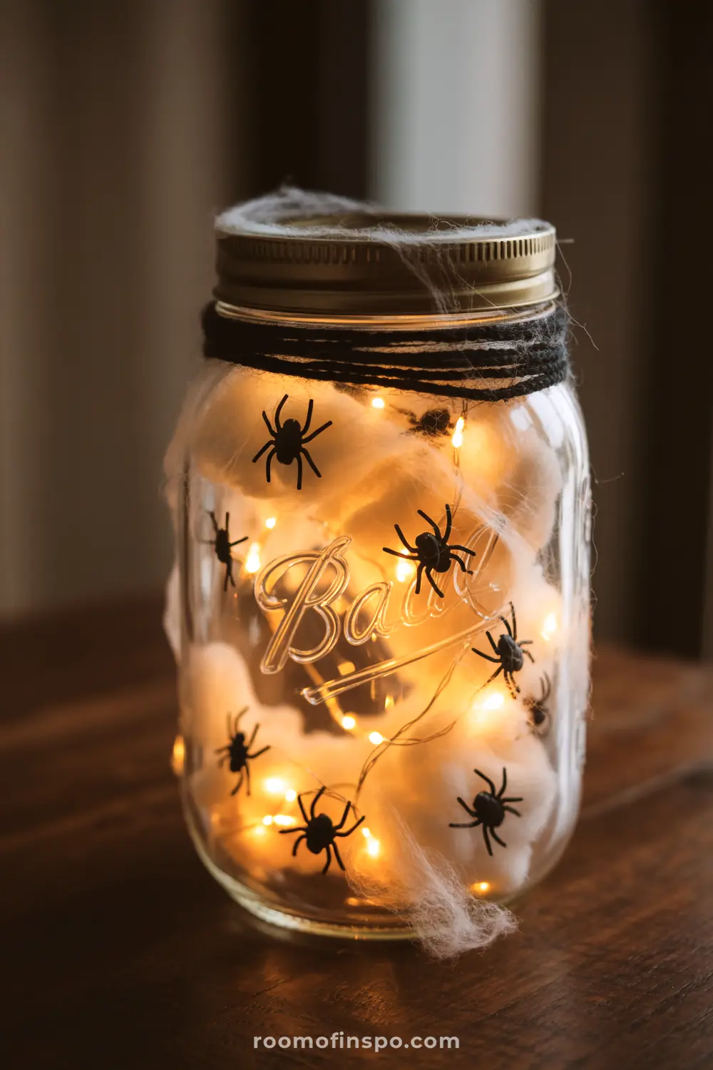 A glowing Halloween centerpiece features a mason jar filled with fairy lights, cotton batting for web effect, and plastic spiders.