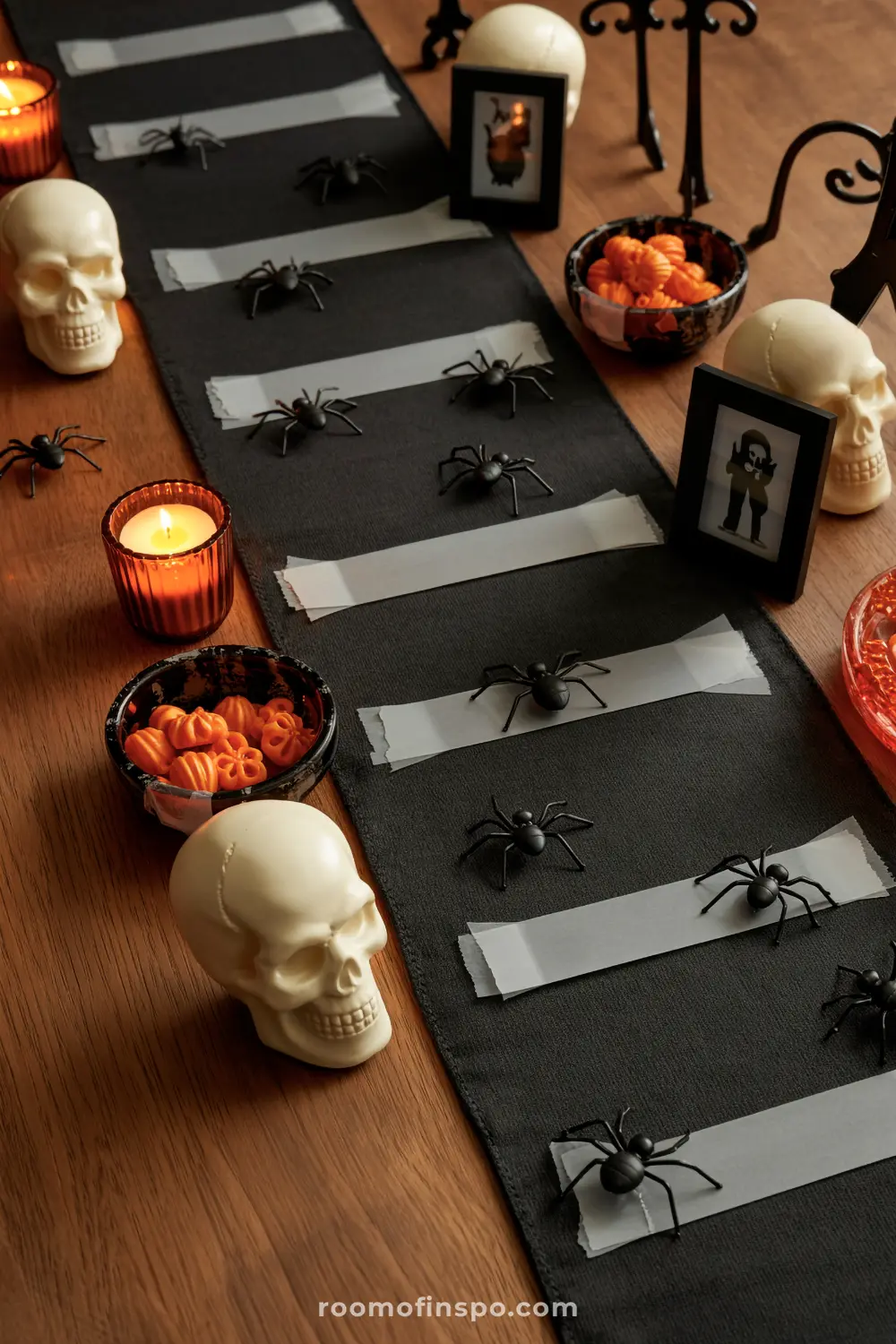 A creepy Halloween centerpiece with skulls, black spiders, and orange candy in bowls, arranged on a dark runner with white tape.