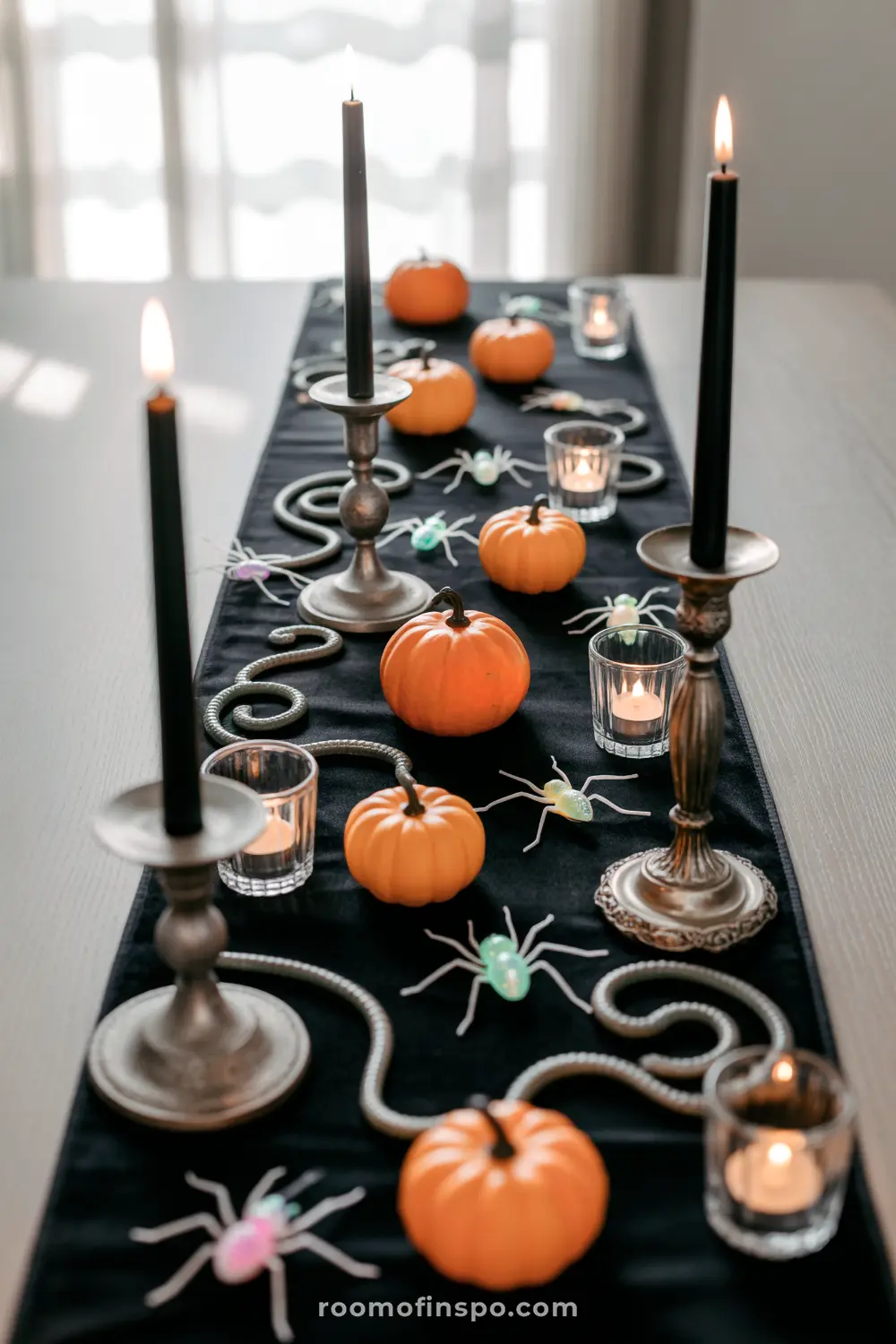 A playful Halloween centerpiece on a black runner featuring orange pumpkins, black candles, colorful spiders, and winding silver snakes.