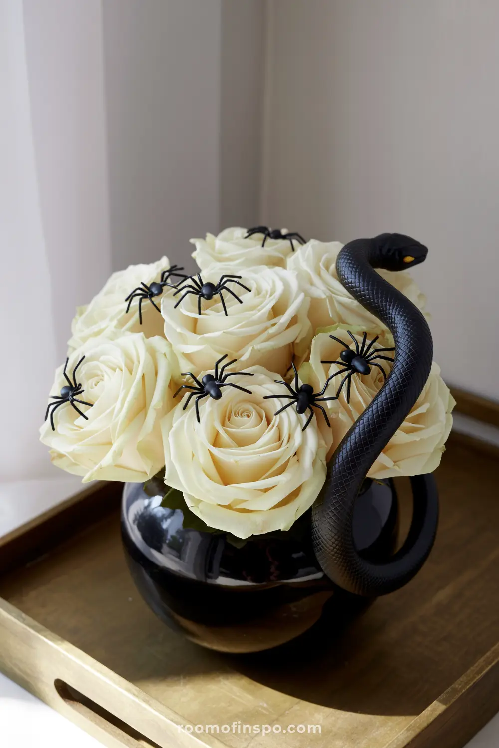 A subtly eerie Halloween centerpiece featuring a black vase filled with white roses, adorned with black spiders and a coiled black snake.