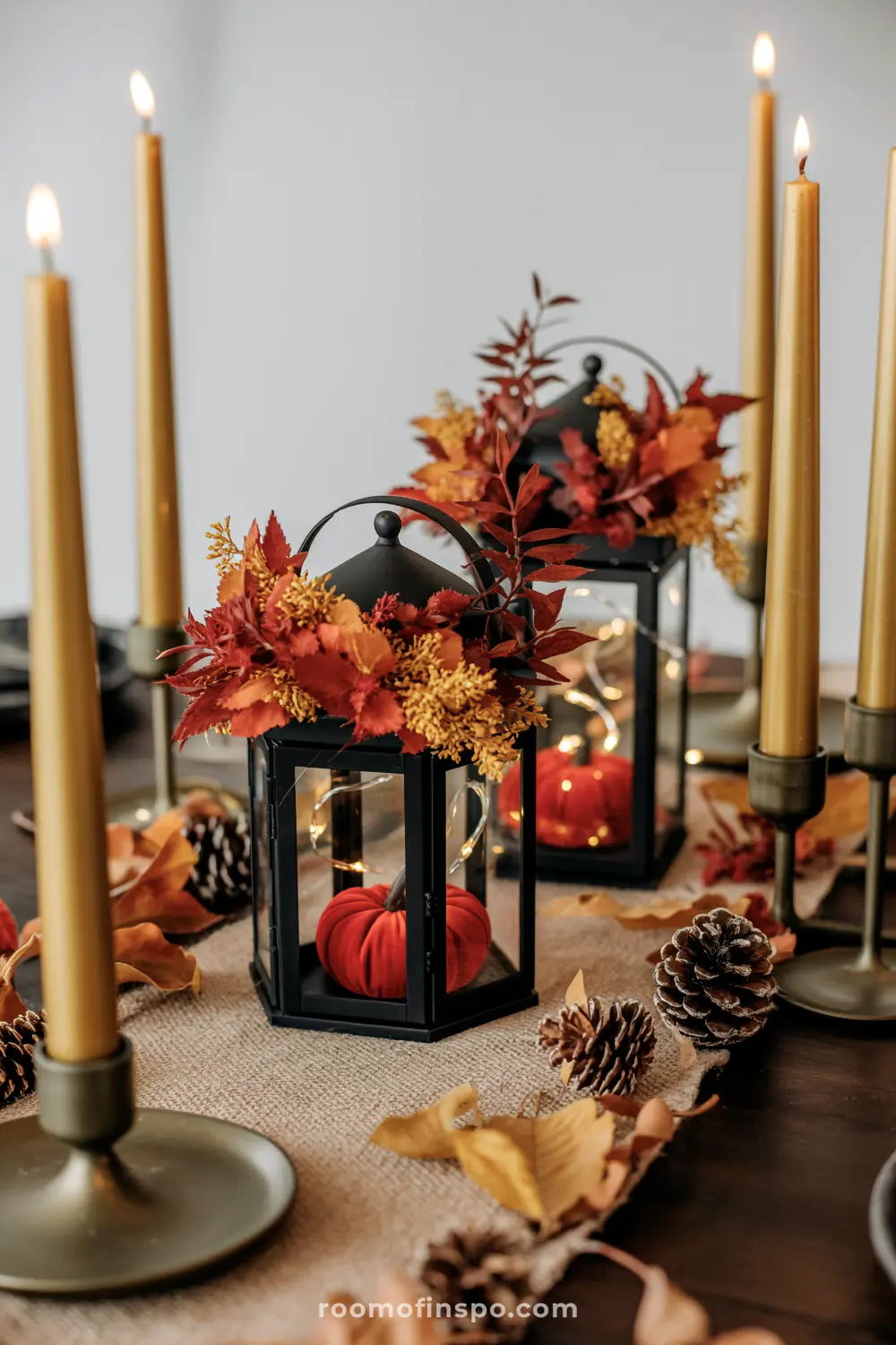 A cozy Halloween centerpiece featuring black lanterns with velvet pumpkins and fall foliage, surrounded by tall gold candles and pinecones.