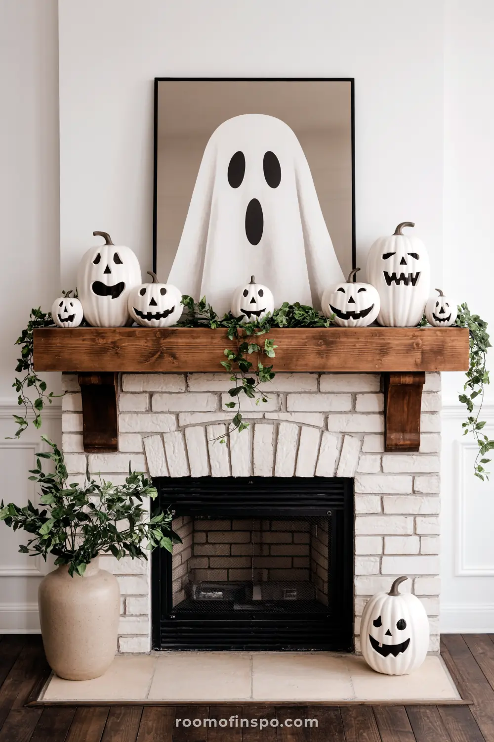 A whimsical Halloween mantel features a ghost artwork, white carved pumpkins, and green garland on a rustic wooden beam over a brick fireplace.