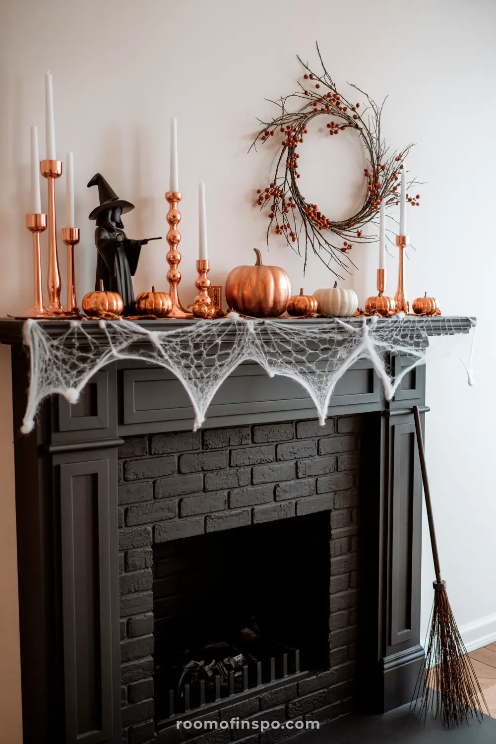 A spooky Halloween mantel on a dark fireplace features white cobwebs, a witch figure, copper-toned pumpkins, candles, and a berry wreath.