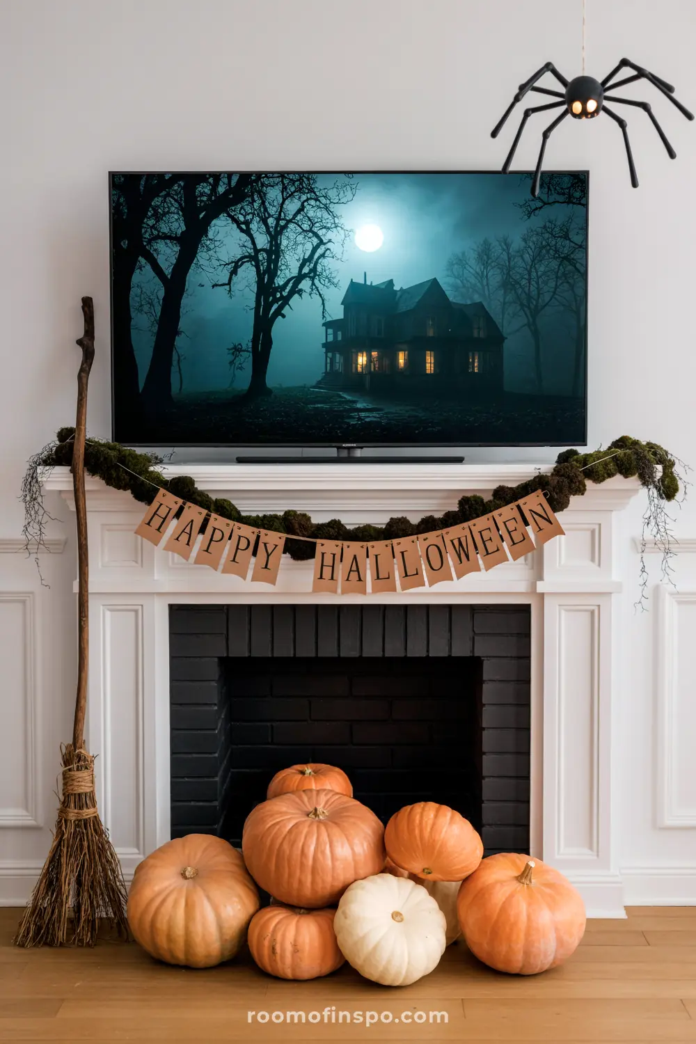 A white mantel is decorated for Halloween with a "Happy Halloween" banner, moss, and various pumpkins. A large spider hangs above the TV.