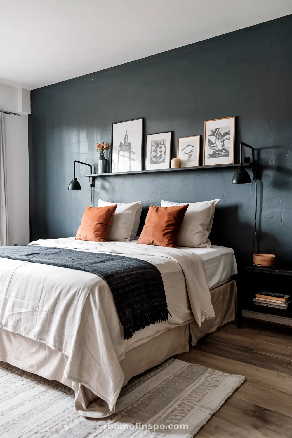 A contemporary bedroom with a headboard-free bed featuring crisp white linens, rust-colored pillows, a dark gray throw blanket, a black shelf displaying framed art, wall-mounted reading lamps, a black nightstand with books and a wooden bowl, and a light-colored textured rug on a wooden floor.