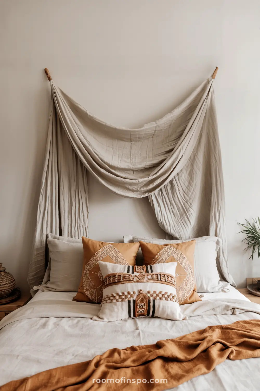 A cozy bedroom with a headboard-free bed featuring white sheets, a brown blanket at the foot, decorative pillows with geometric and patterned designs, beige fabric draped above the bed for a canopy effect, and a small plant on a bedside table.