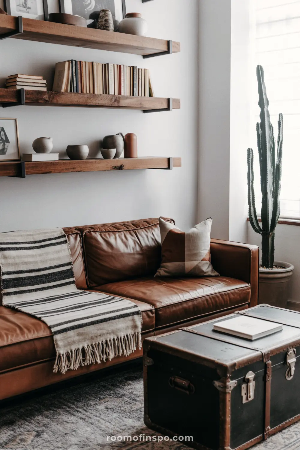 A masculine living room with a brown leather sofa, open shelving with books, and a trunk for a coffee table.