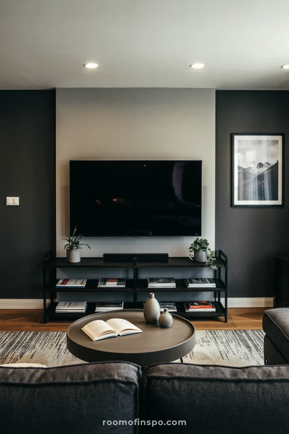 A gray and black masculine living room with a large TV and a black, round coffee table with vases on it.