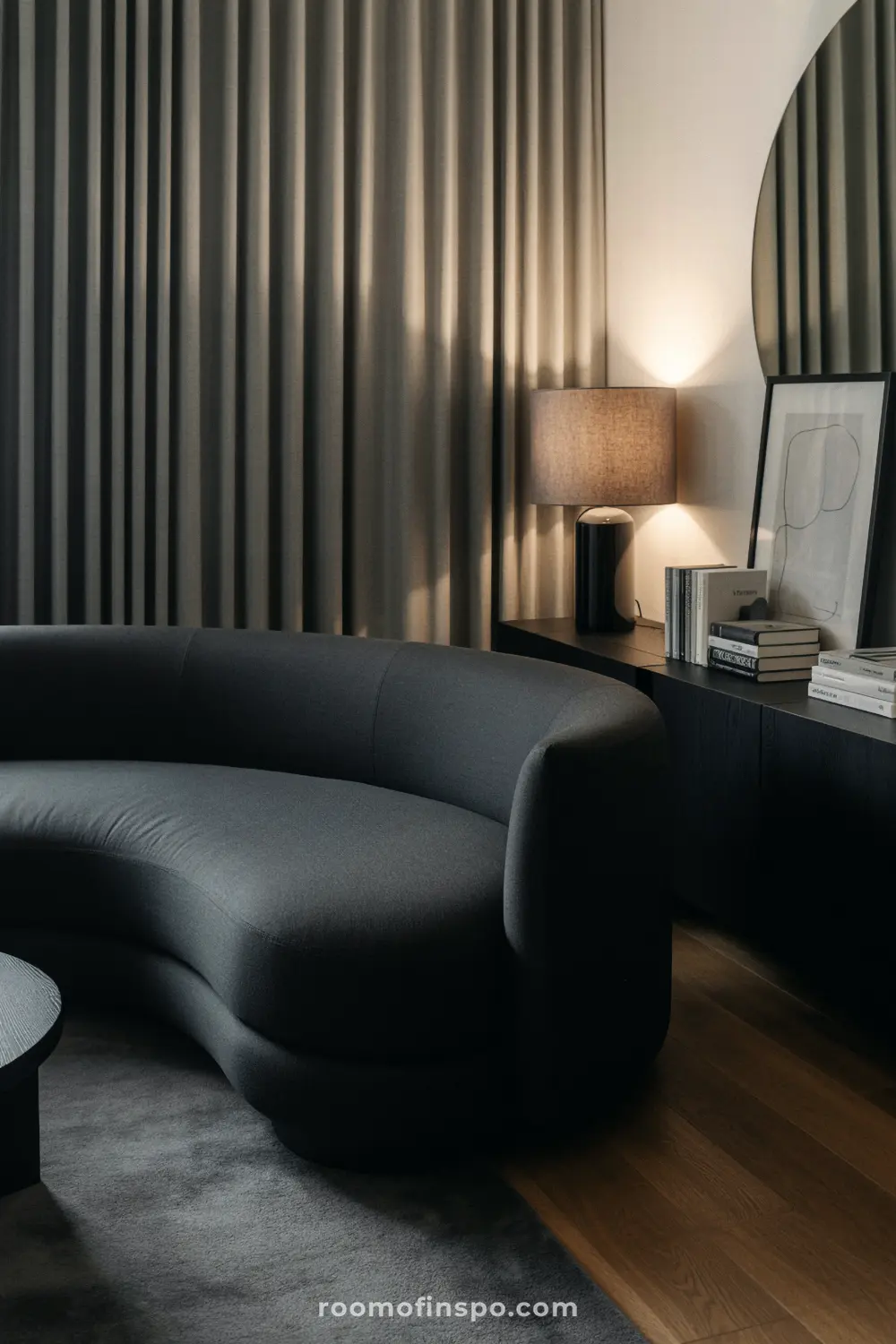 A man's living room with a unique curved gray sofa and a circular black table on a gray rug.