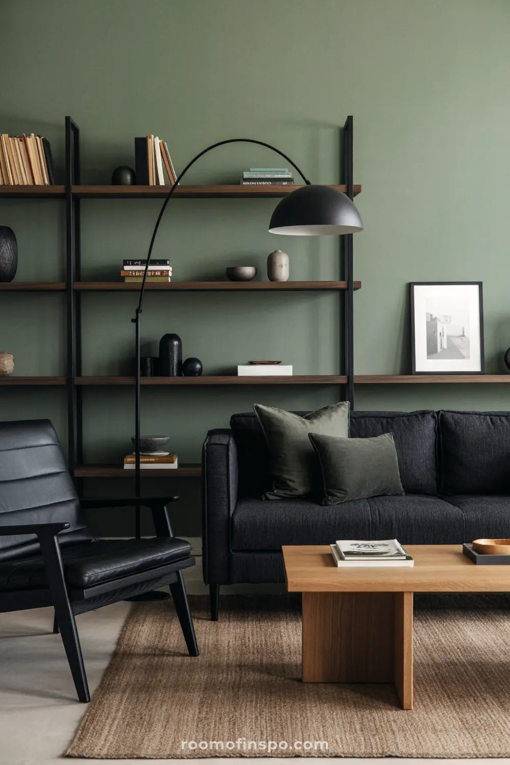 A man's living room with a sage green accent wall, a black sofa, a black leather armchair, and a tall arched floor lamp.