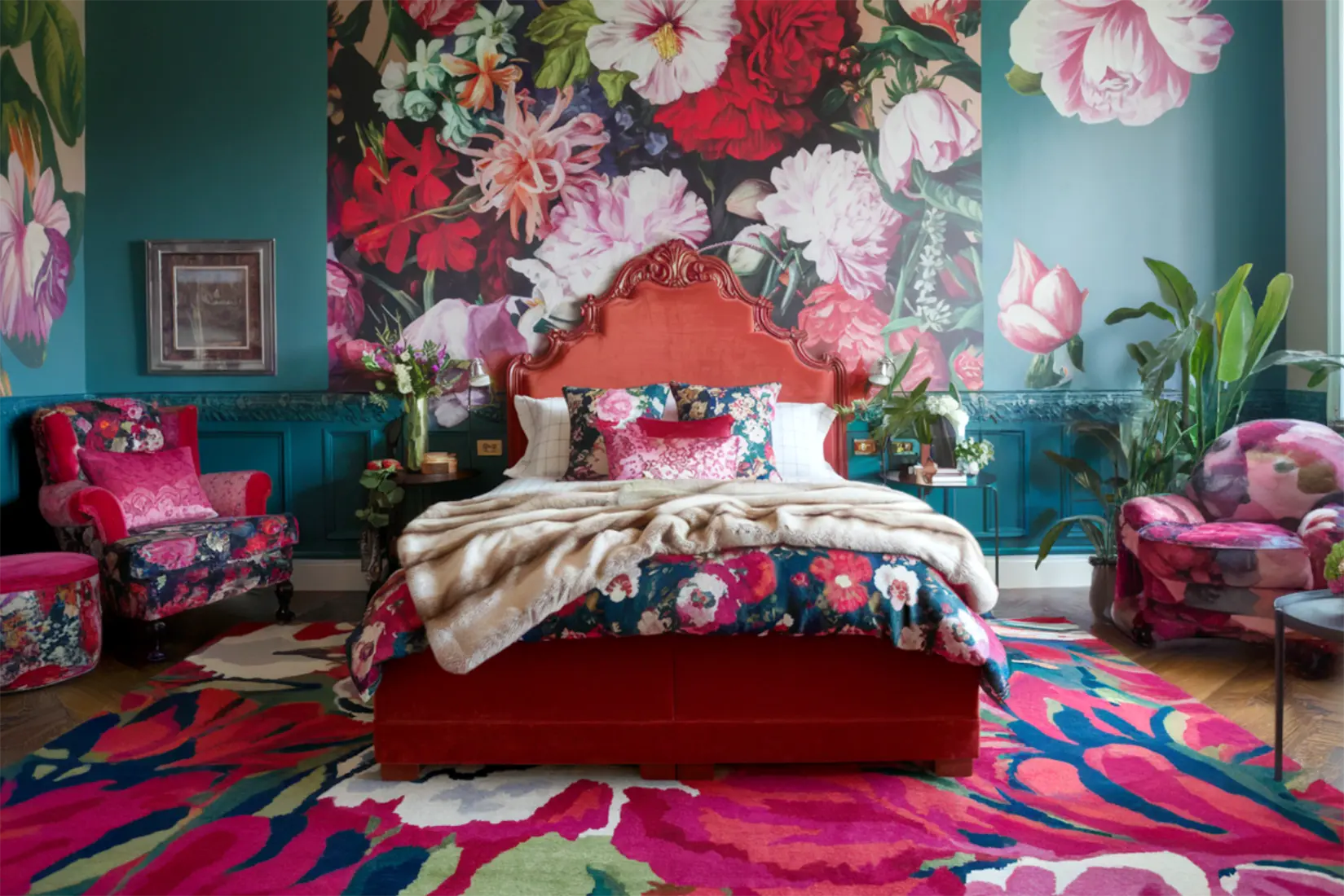 A vibrant and eclectic bedroom with a large floral mural on the wall, a plush red velvet bed, and patterned armchairs and rugs.