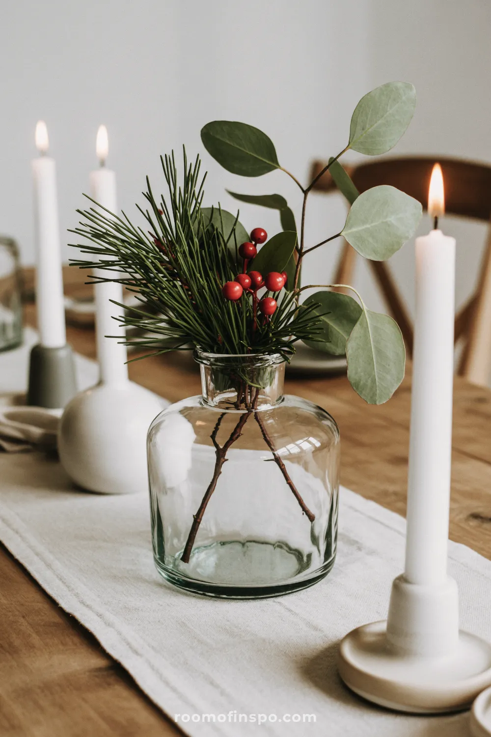 A clear vase holds pine, eucalyptus, and red berries, creating a minimalist Christmas centerpiece with candles.