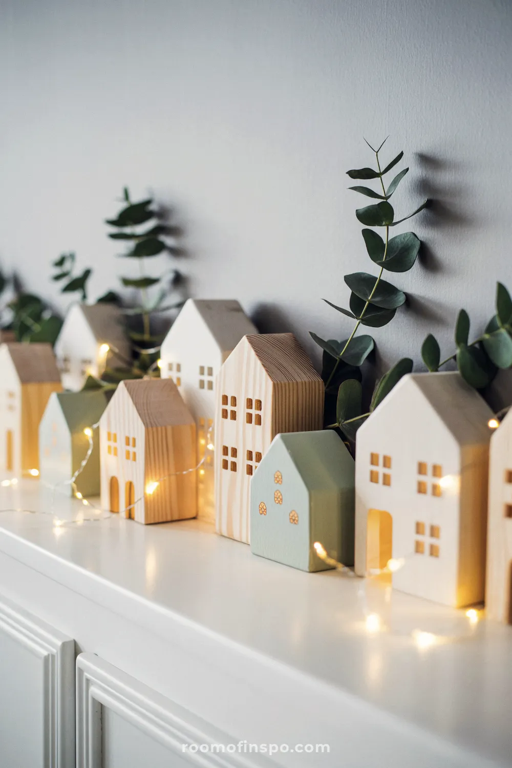 A shelf holds a row of light wooden and white miniature houses with string lights for a minimalist Christmas scene.