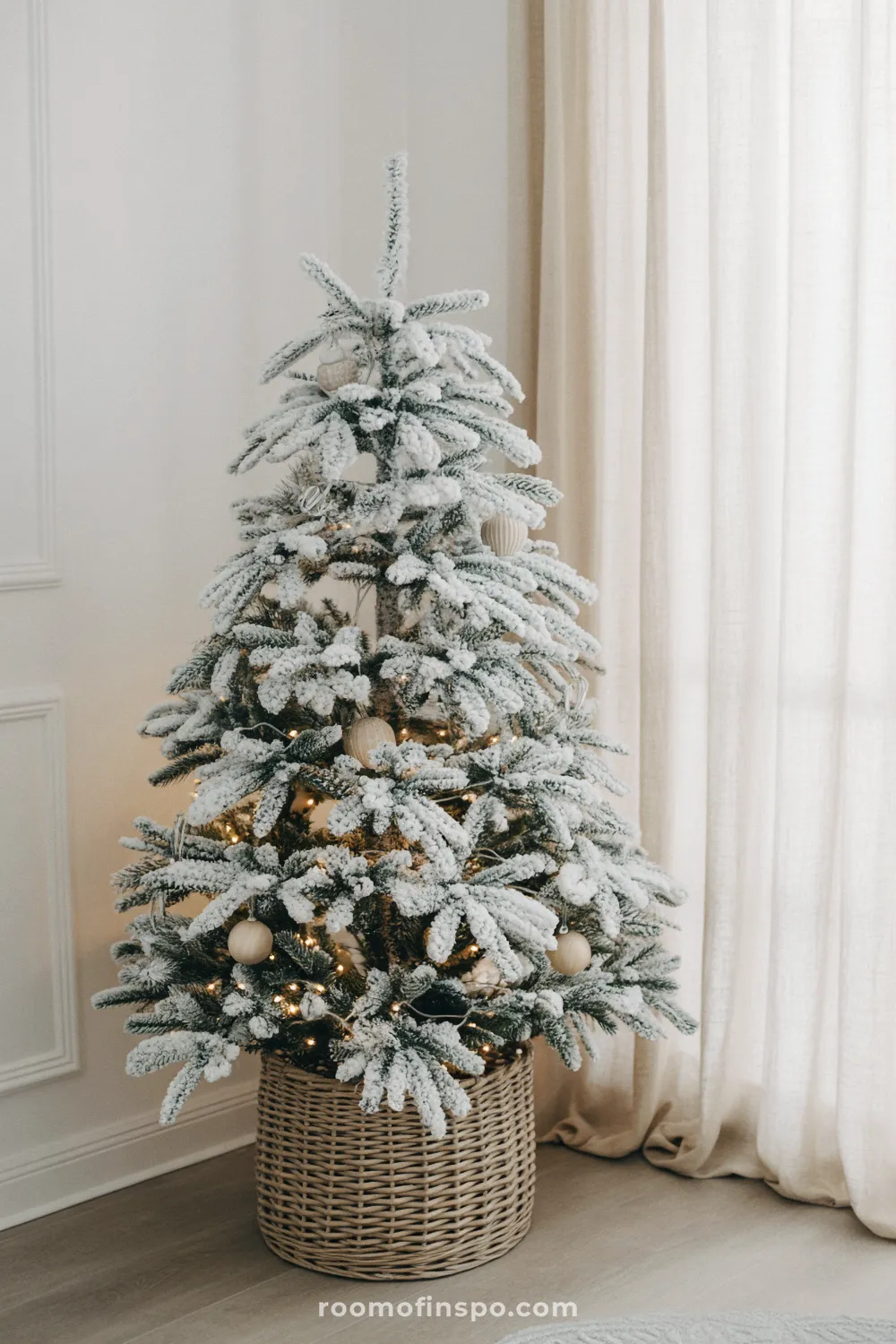 A snow-dusted, unadorned tree in a wicker basket stands by sheer curtains for a minimalist Christmas look.