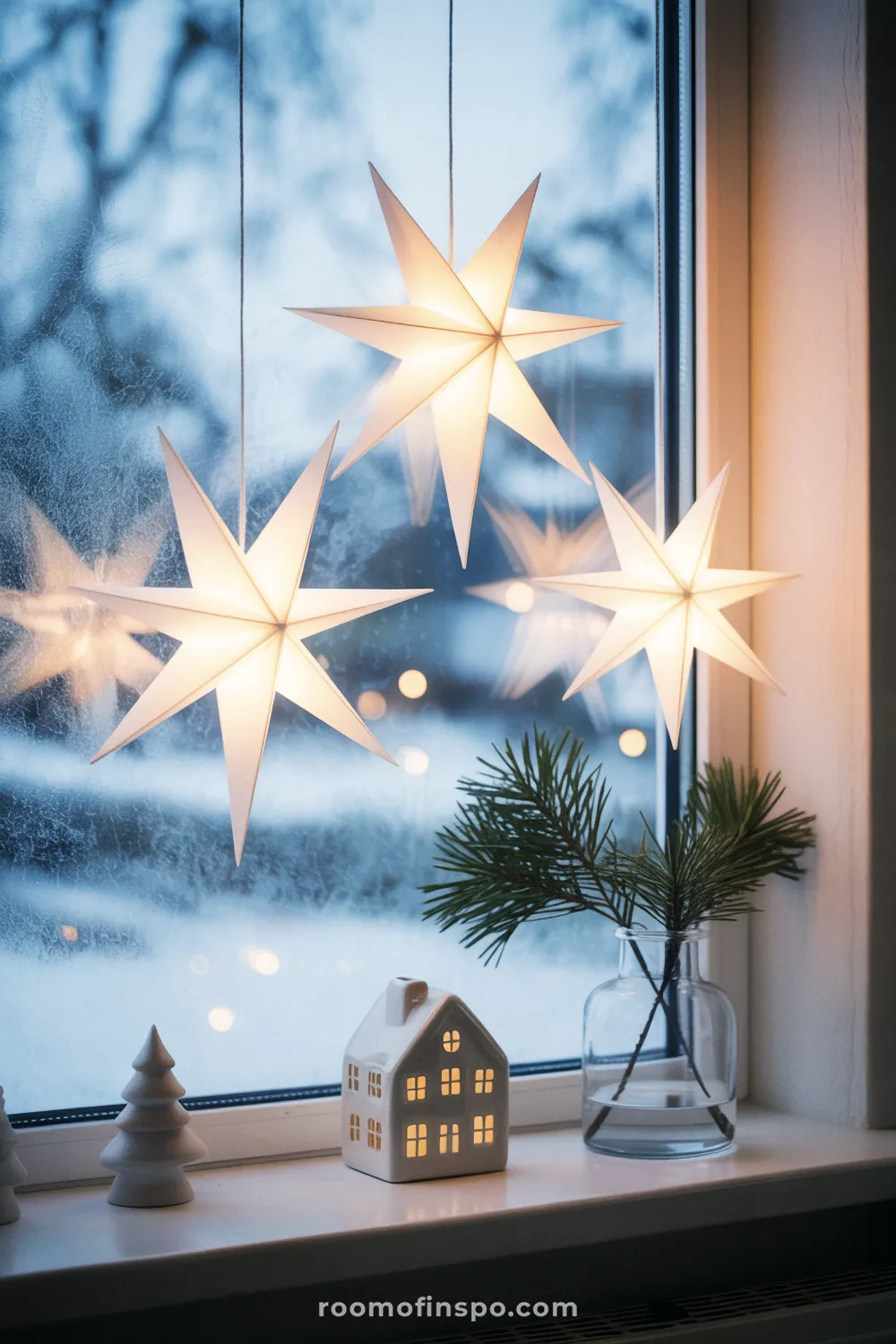 Three illuminated paper star lanterns hang in a frosted window with a tiny lit house for a minimalist Christmas scene.