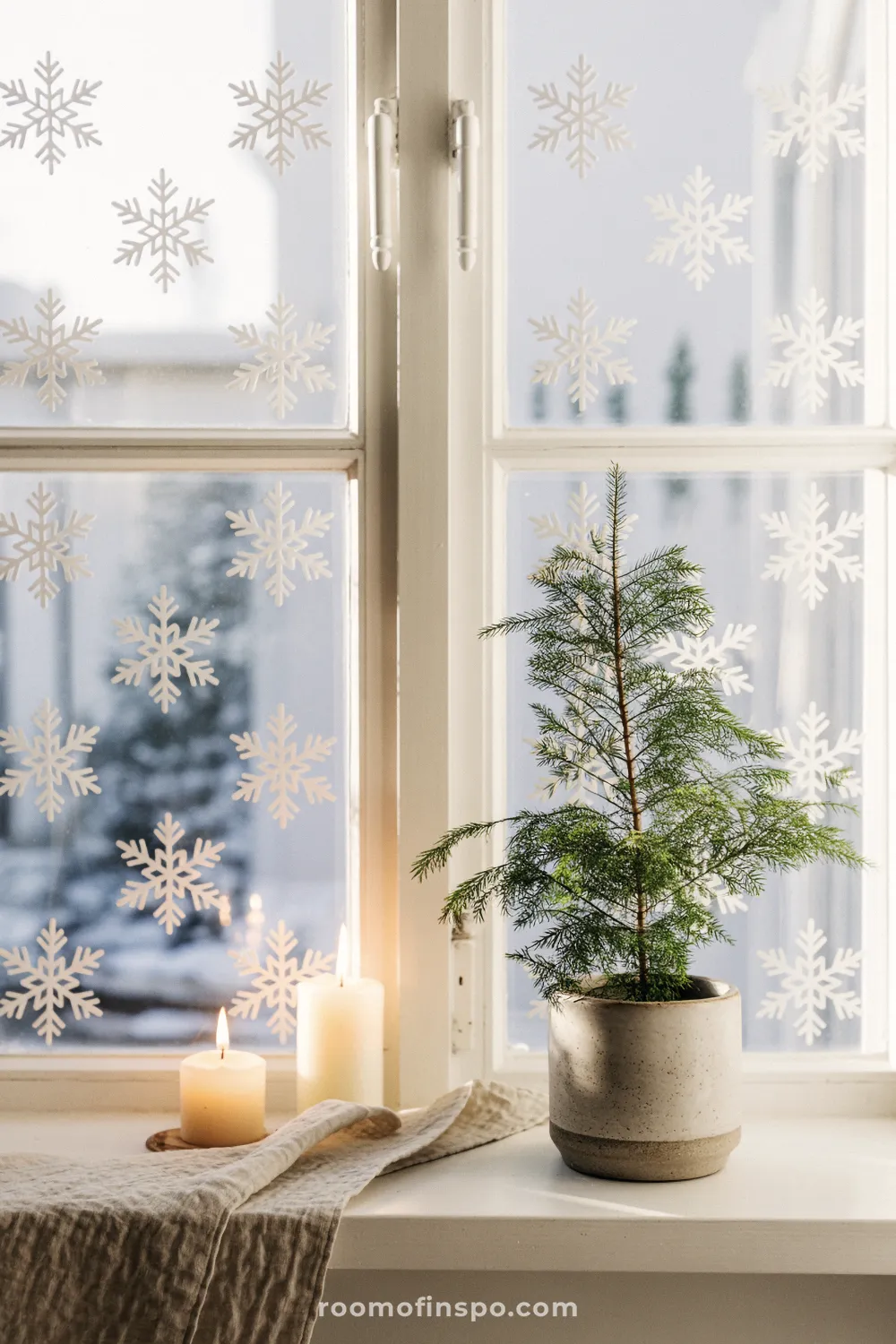 A small potted tree and candles are placed on a windowsill decorated with snowflake decals for a minimalist Christmas feel.
