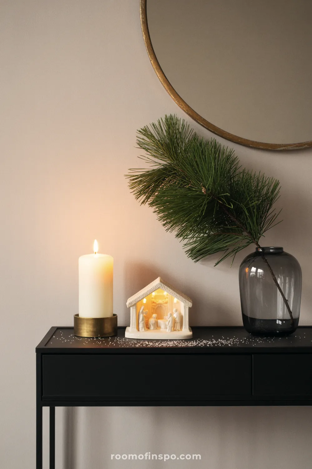 A black console table is decorated with a lit nativity, a candle, and a pine sprig for minimalist Christmas decor.