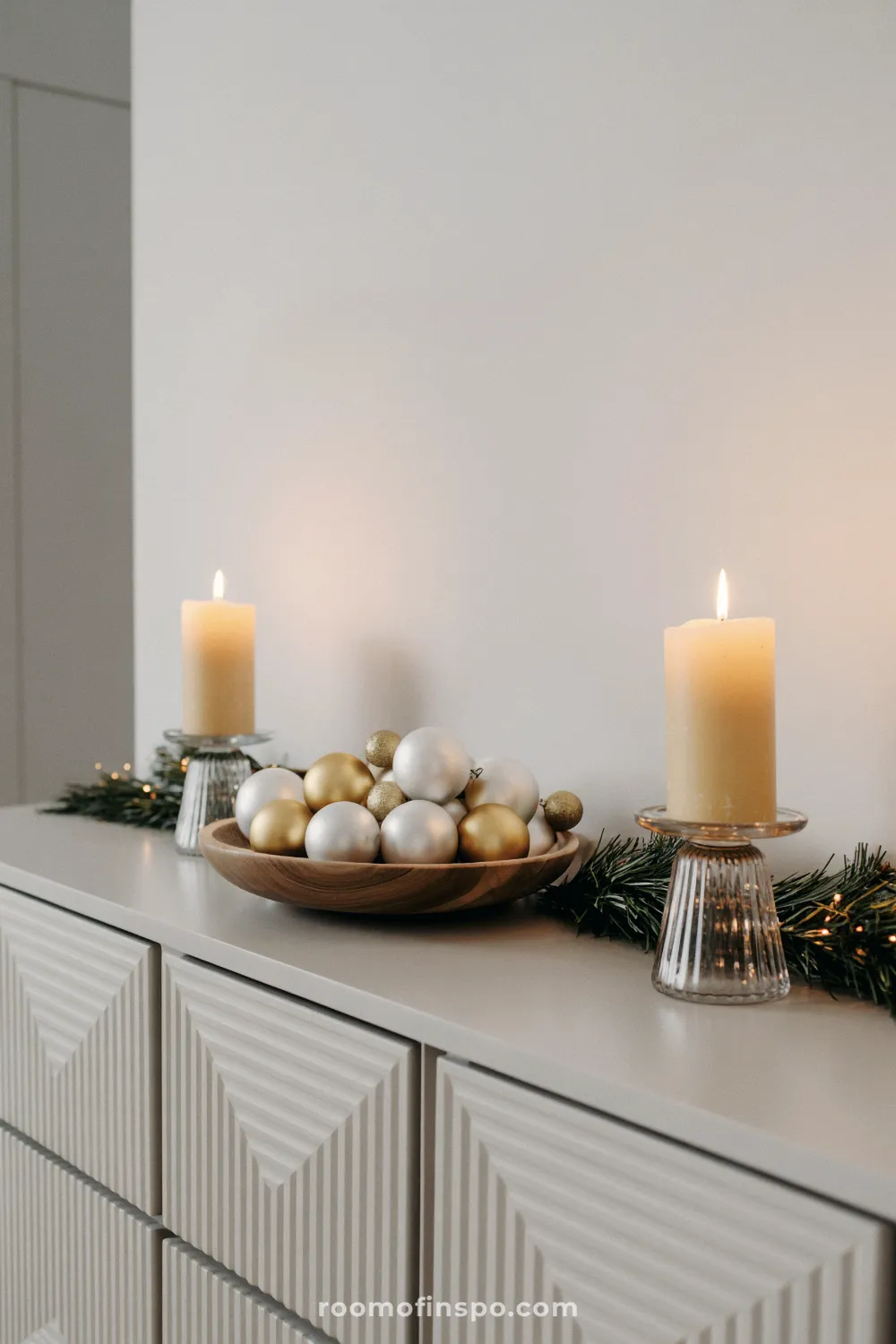 A textured gray cabinet is topped with gold and silver ornaments in a bowl between two candles for a minimalist Christmas look.