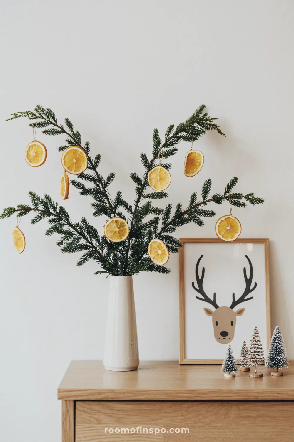 A wooden console table holds a vase with pine branches and dried orange slices for minimalist Christmas wall art.