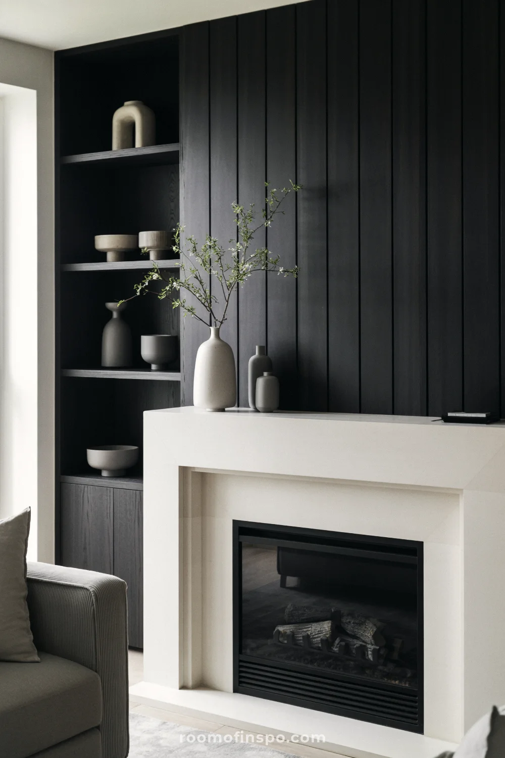 Minimalist living room with white concrete fireplace, charcoal paneling, neutral ceramics, gray armchair, and bright window light.