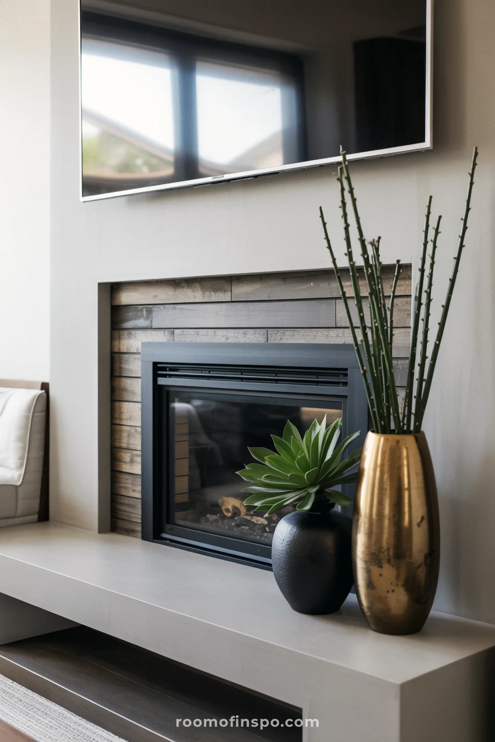 Modern fireplace with black-framed opening, weathered gray wood surround, concrete mantel, gold and black vases, and TV above.