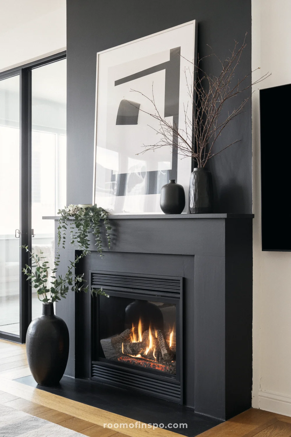 Dark minimalist living room with black gas fireplace, burning flame, ceramic vases, abstract art above, oak floors, and window.