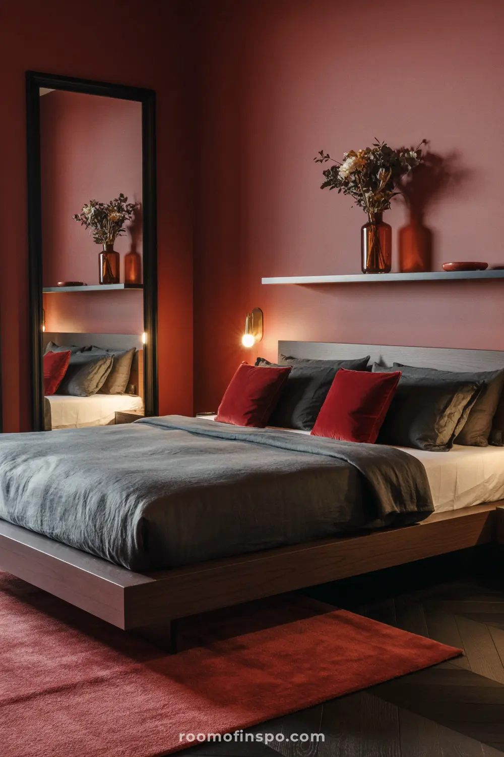 A moody bedroom with dark gray bedding and red accent pillows, featuring a large mirror and floating shelf on a deep red wall.