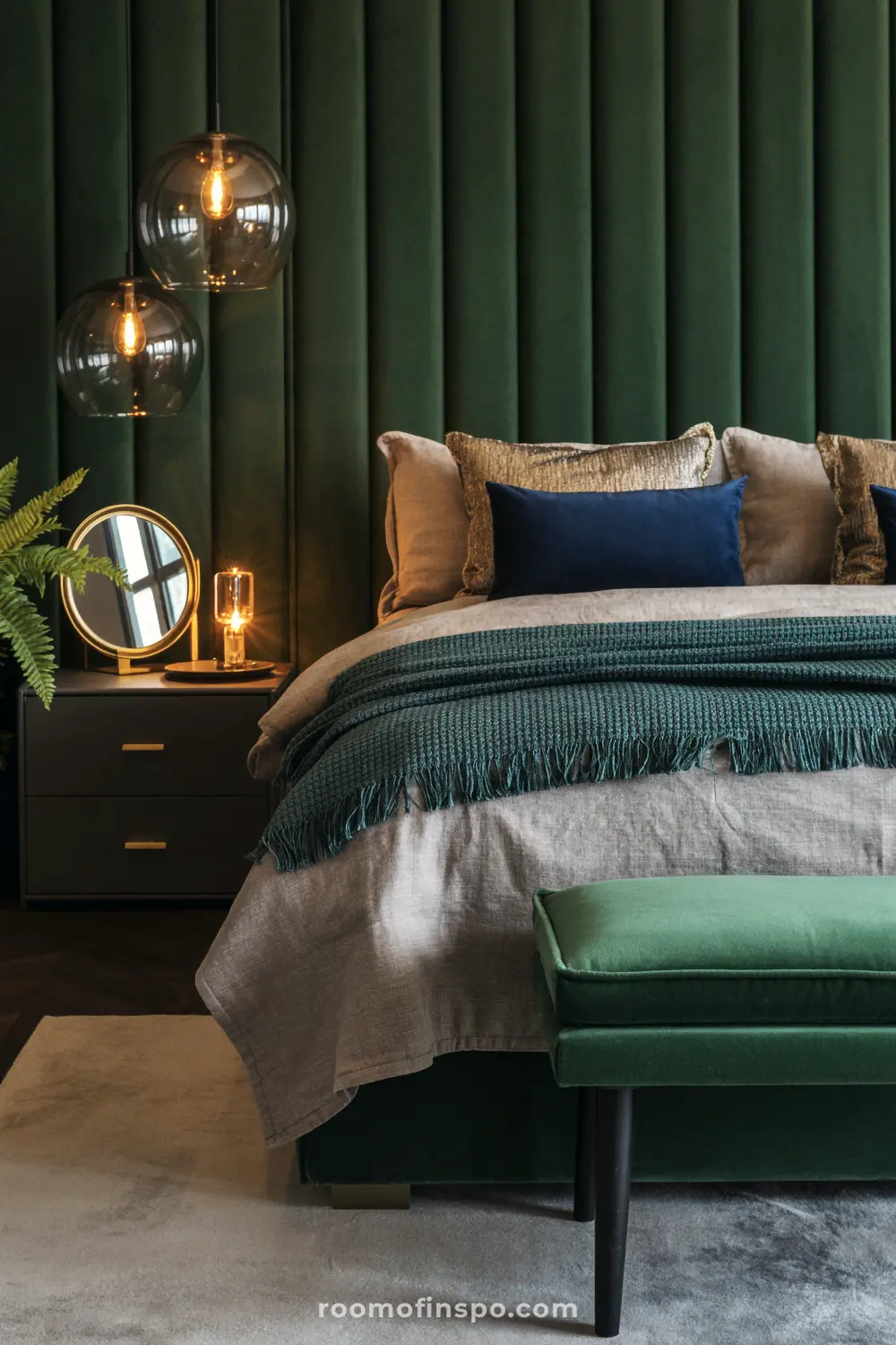 A luxurious and dark bedroom featuring a plush, forest-green velvet headboard and matching bench, with modern pendant lights and a gold-accented nightstand.