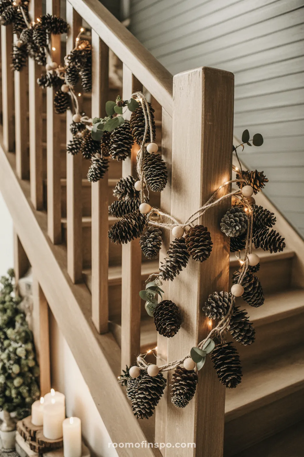 A light wood staircase banister is wrapped with a beautiful natural garland of pinecones, wooden beads, and lights for Christmas.