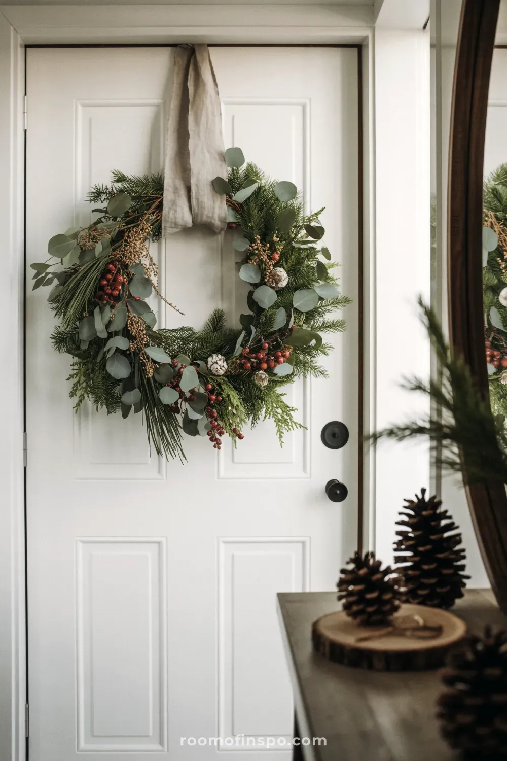 A full wreath of mixed greenery, berries, and cones hangs on a white door for a minimalist Christmas entryway.