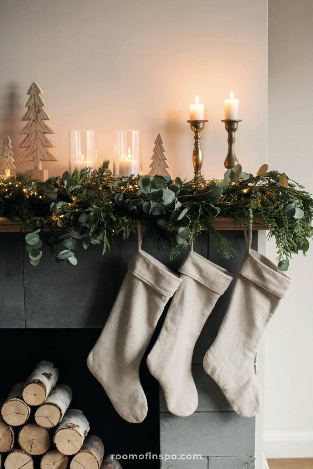 Three simple linen stockings hang below a natural garland on a fireplace mantel decorated for Christmas with candles and wooden trees.