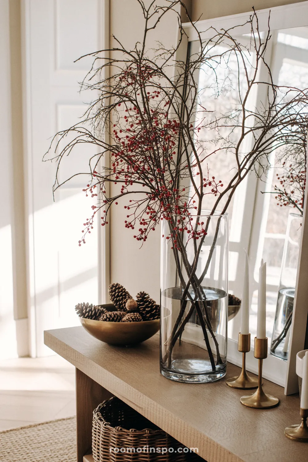 A vase holds natural winter branches with red berries and a bowl of pinecones on a console table for Christmas cheer.