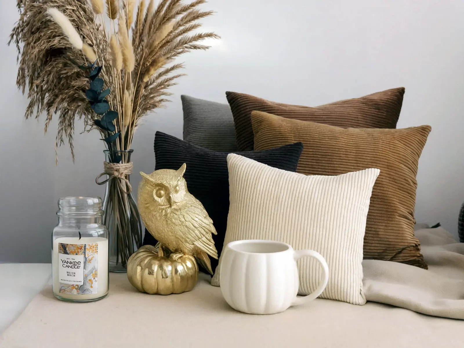 Neutral fall decor items including a vase with dried grasses, a gold owl on a pumpkin, a candle, a mug, and corduroy pillows in earthy tones.