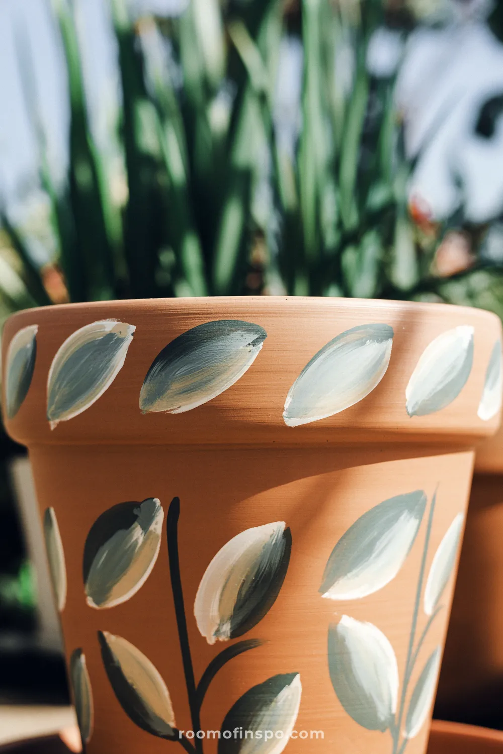 Close-up of a terracotta pot painted with muted green vine leaves against a blurred garden background.