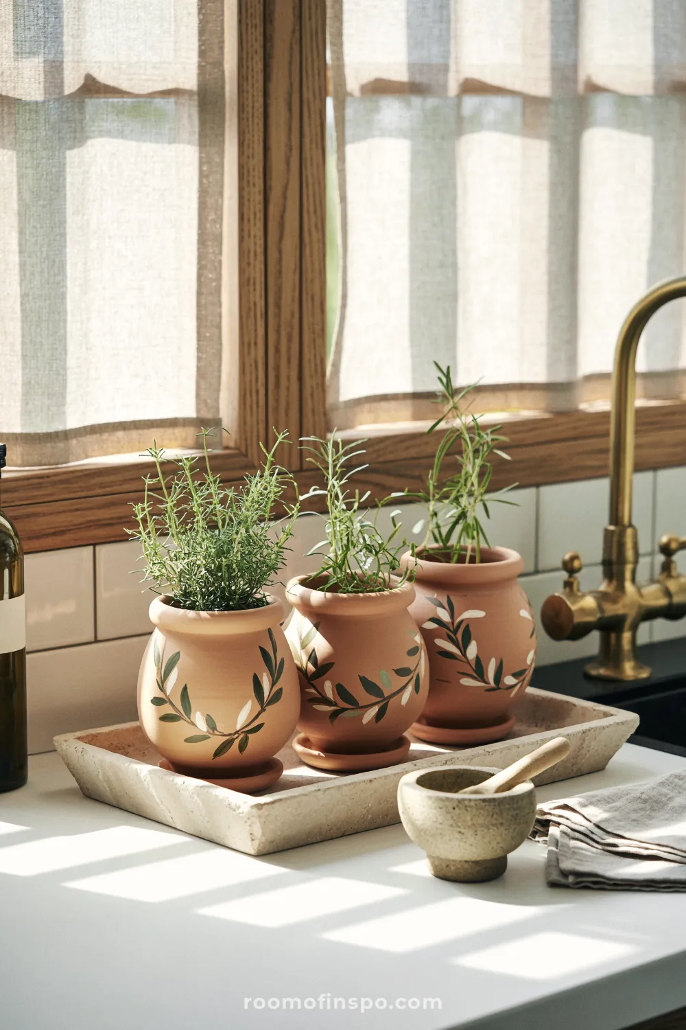 Three vine-painted terracotta herb pots on a kitchen windowsill beside a brass faucet and linen curtain.