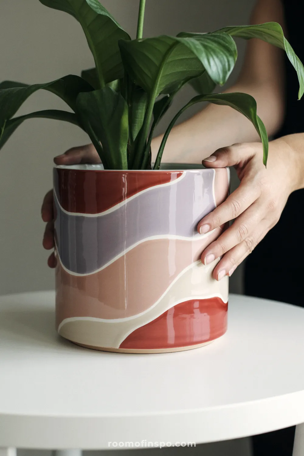 Modern ceramic pot with blush and lavender wavy pattern on a white tabletop with a leafy plant.