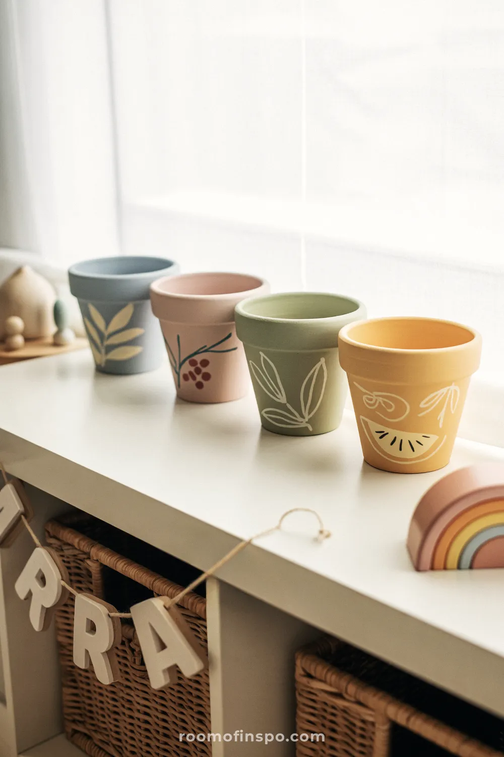 Four pastel-painted terracotta pots on a white shelf with woven bin and small wooden toys nearby.