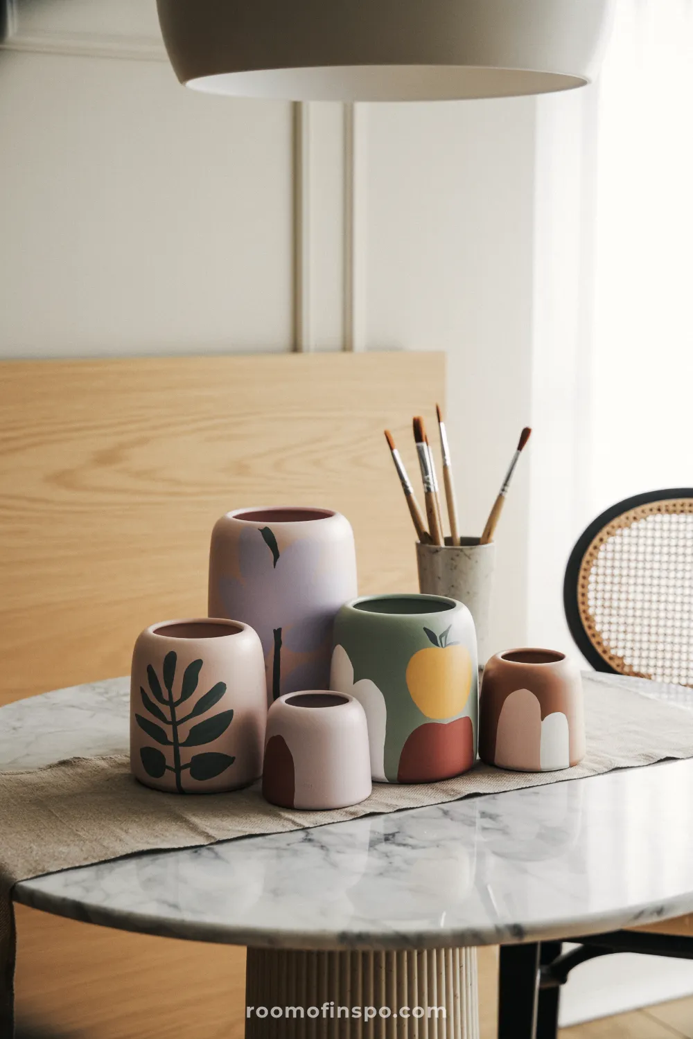 Assorted pastel botanical pots grouped on a marble bistro table with linen runner and cane chair in background.