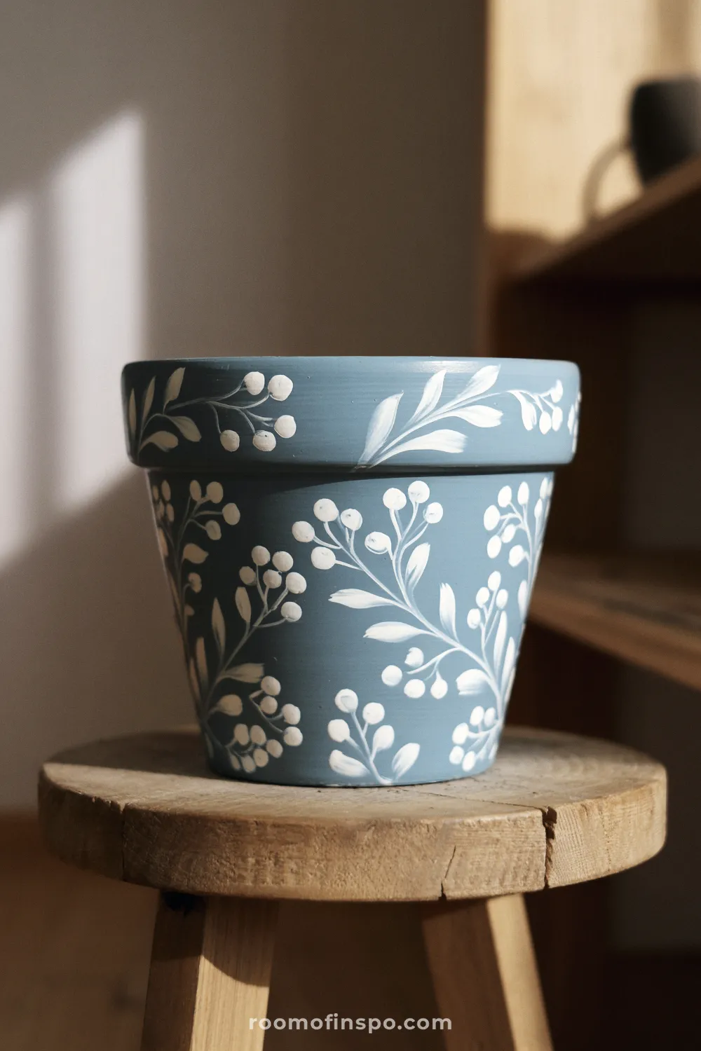 Dusty blue terracotta pot with white floral pattern on a wooden stool in warm indoor light.