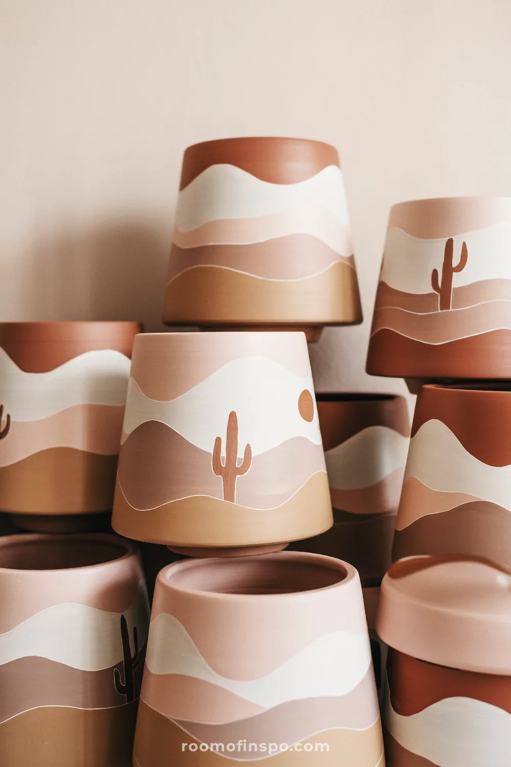 Set of terracotta pots painted with layered desert landscapes, cactus silhouettes, and sun shapes against a neutral wall.