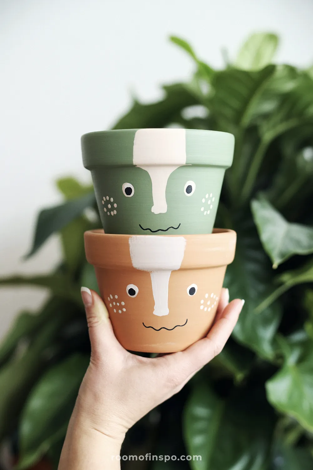 Two stacked pots with simple face designs, one sage painted and one natural terracotta, held outdoors.