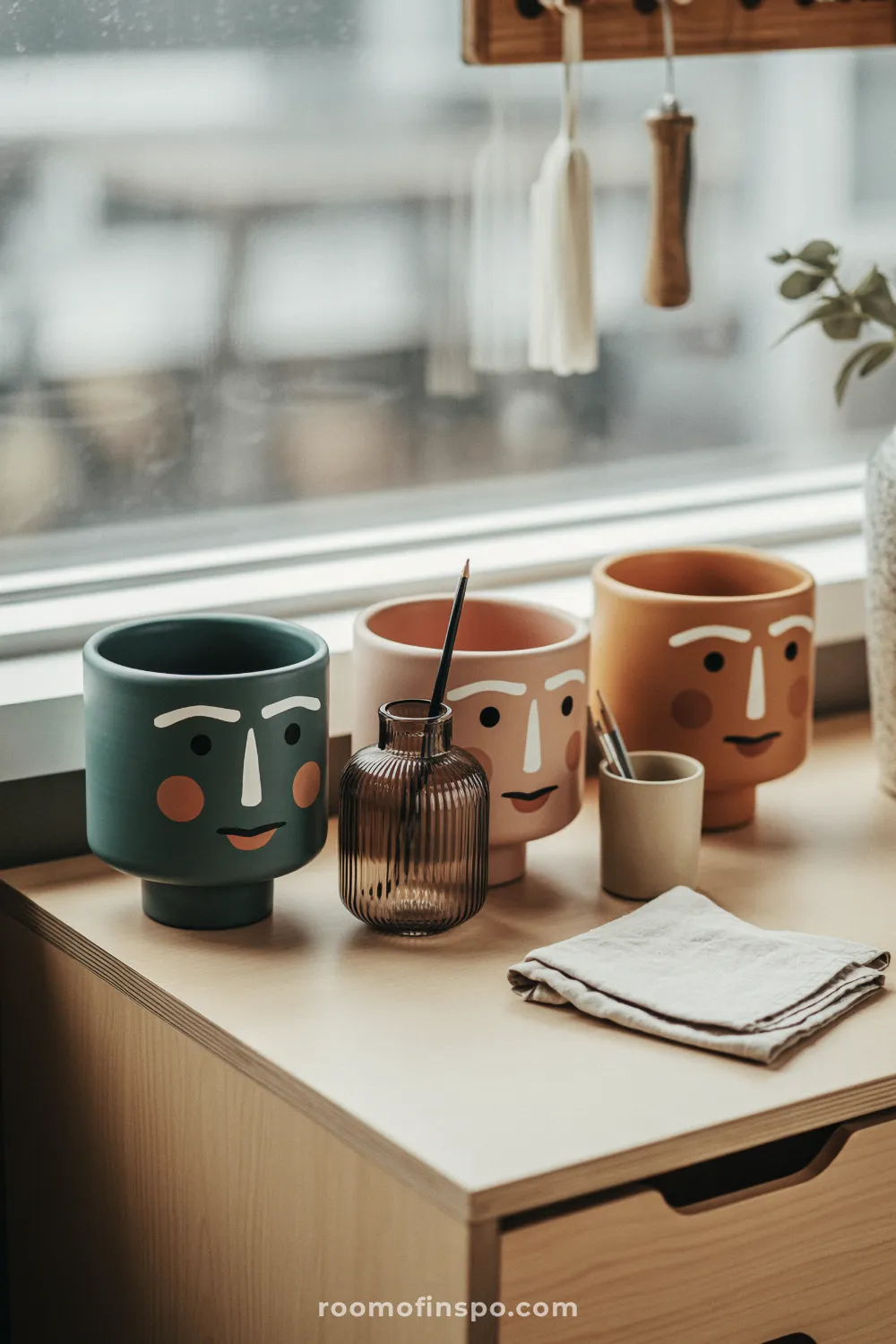 Six small pastel pots with simple painted faces lined up on a white shelf with succulents.
