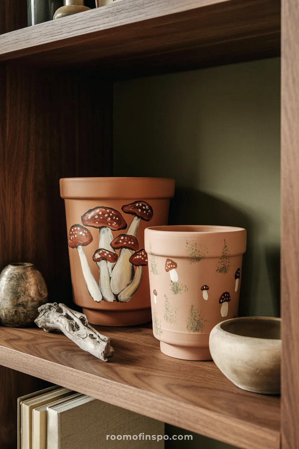 Two mushroom-painted terracotta pots on a walnut shelf with driftwood and stoneware bowl under warm lamp light.